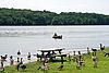 A lake with a canoe and forested far shore, a flock of Canadian geese and picnic table are on the grassy near shore