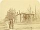 Sepia-toned 1890s photograph showing two men in late 19th-century clothing standing on the street corner in front of a brick house