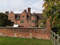 A photograph of a large, three-storey brick house, set well back behind a garden wall.
