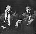 Two men in business suits sit in theatre seats amiably discussing what they are watching