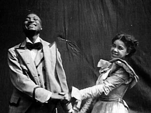 A black man in a suit smiles and holds the hand of a black woman, who looks at the camera and smiles. Both are dressed in clothes from 1898.