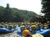 Dozens of people wearing yellow life preservers and helmets in several blue inflatable rafts float down a river