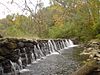 Several streams of water fall over a dam made of large rocks under autumn leaves