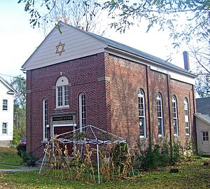 A brick building with a small gold-on-black sign saying "Congregation Beth David" above the door, round-arched windows and a small structure in front made of withered cornstalks on a metal frame