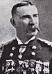Head and shoulders of a white man with a drooping mustache, wearing a military jacket with shoulder boards, one medal at the neck, and a row of medals across the entire chest.