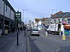 Street scene showing shops on left and right, with cars and vans on road. On the left hand pavement is a sign saying welcome to Keynsham high street.