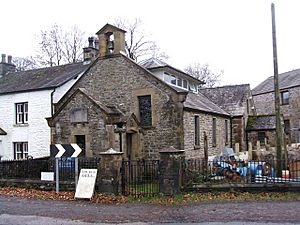 A small stone church with a porch and a bellcote in the foreground, and the body of the church, on which is a glazed lantern on the roof, receding into the background