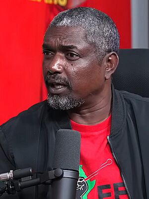 A man seated and behind a microphone, wearing a black blazer and red t-shirt