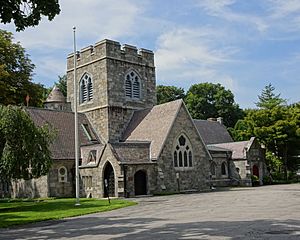 A stone building with a pointed roof and stained glass windows. In its rear is a square tower with crenelated parapet.