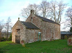 A small bare stone building seen from an angle, a lower section with a doorway at the front, and a larger, higher section with a bellcote at the back