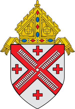 Shield topped by a mitre, featuring a silver field divided per red saltire, four red crosses within the four quarters, and a silver wind mill on the saltire