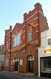 A tall building with deep red brickwork and yellowish stone dressings. There are two identical entrances: wooden doors in recessed stone doorways with prominent yellow keystones.  A window to their left is in a similar recess.  Two pediments flank a stone frieze with the words "THE SALVATION ARMY".  The upper floor has two rectangular windows and a central arched window with another keystone.