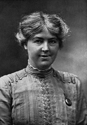 Monochrome photograph portrait of a woman in her twenties, shown from the bust upward, the woman wearing a vertical-patterned blouse decorated by a row of buttons between the shoulders and the closed collar, her face directly forward gazing at the viewer, her cheeks prominent and fleshy, the mouth slightly opened in a tight smile, the coarse, sun-bleached sandy-coloured hair parted in the middle, extending to the ears in an overall loose wave with flyaway strands