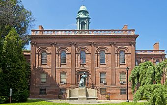 A two-story brown stone building with ornate classical detailing in a park setting. It has a green cupola on top and a green statue of a man in front.
