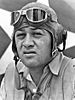 Head of a squinting man wearing a shirt unbuttoned at the collar and a cloth aviator's cap with headphones built into the ear flaps, an unbuckled chin strap, and goggles pushed up onto his forehead.