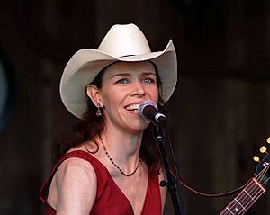 A slender, middle-aged woman with long brown hair plays guitar and sings into a microphone. She wears a cowboy hat and a red dress.