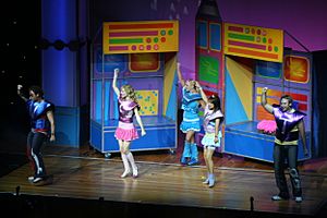 A distant stage shot of five performers, all are in bright clothes and have their right hand raised in the air. All have headset microphones, most are facing to their right and dancing. Behind them are movable sections, each is brightly coloured.