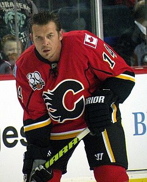 An ice hockey player stands partially crouched, leaning on his stick. He has short black hair and is not wearing a helmet. He is wearing a red uniform with a large black C on his chest.