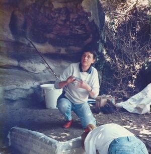 Colour photograph of Dr Val Attenbrow, crouched in a sandstone rockshelter, kneeling in front of an open excavation with another archaeologist working within the square.