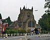 A cruciform church with a broad central tower; this is crenellated and has prominent buttresses; in front is a lych gate and a white railing; in the foreground is a lamppost and a road with traffic islands