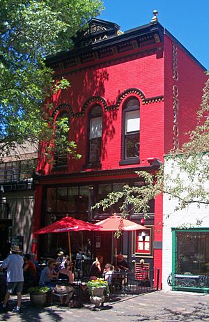 A narrow three-story red brick building with ornate black trim, connected to lower buildings on either side, seen from its right, with tree branches entering the frame from either side. The words "Red Onion" are in vertical gold lettering on the right side. In front are tables with red umbrellas and people seated around them.