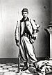 Full-body portrait of a white soldier with a rifle and bayonet by his side, standing on a crumpled flag. He is wearing baggy pants tucked into his boots, a short, decorative jacket buttoned at the top only, and a slouch cap.