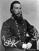 Black and white photo shows a full bearded man, seated with his left hand holding his right hand. He wears a military uniform with two rows of buttons and frogging on the sleeves.