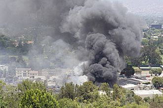 A large plume of gray smoke rises from a complex of buildings in a wooded area, seen from slightly above