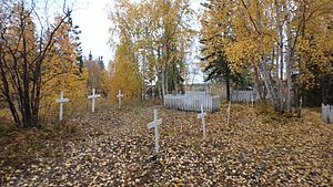 Two parallel rows of three white crosses, one at left and one in the centre, on a mostly dirt terrain covered with autumn leaf litter. The background is dominated by trees with dark yellow leaves, and one leafless bush with a dark trunks is at left behind the first cross.