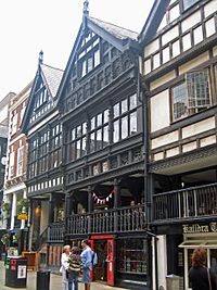 A black-and-white building with two gables and three stories; a shop and a stairway on the ground floor, wooden railings at the edge of the Row, large windows and panels in the storeys above