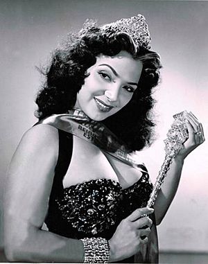 María Victoria smiling while wearing a black gown, a crown, and holding what it looks to be a sceptre