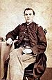 Young white man sitting on a chair with his legs crossed and his right arm resting on a table beside him. He is wearing light colored pants and a military jacket buttoned at the top only with a vest underneath.