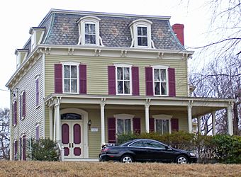 A three-story wooden house, light brown with purple shutters and a mansard roof shingled in fish-scale slate with dormer windows. There is a porch on the first story and a black car in front.