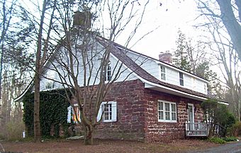 A house built of red stone house with a curved, sloping roof and white wooden top, seen from the side. It has brick chimneys at either end and some ivy on the visible side.