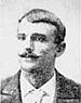 Head of a white man with neatly combed hair and handlebar mustache wearing a suit and tie.