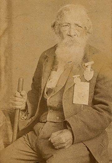 The picture is a sepia toned photograph that shows an African-American man in his eighties wearing a suit adorned with war medals and holding a cane.