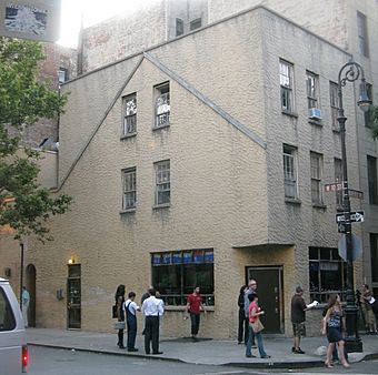 A gray stucco-faced building seen from across a street, with a brown and white sign saying "W. 10th ST" and people on the sidewalk in front.