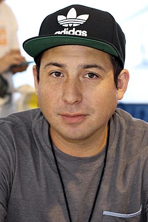 A portrait of Tommy Orange, wearing a hat, grey t-shirt. He is smiling towards the camera