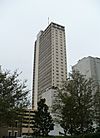 Distant ground-level view of a 35-story skyscraper with a rectangular cross section; the lateral side of the building has a concrete facade, whereas the anterior side has an almost complete glass facade that is light gray in color. A concrete shaft extends up the anterior side and projects beyond the roofline of the building. An antenna mast extends from the building's roof.