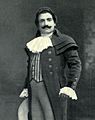  Man, with dark hair and a curling moustache, standing in a posed position. He is wearing a long coat, with lace at the throat and cuffs.