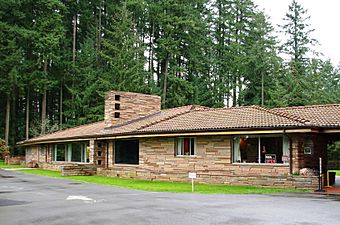 Brown one-story  modern flagstone building, parking lot in front and fir trees behind it.