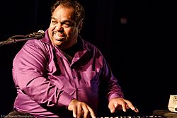 Daryl Davis wearing a purple button-down shirt, playing a piano, turned right-of-camera, towards a microphone