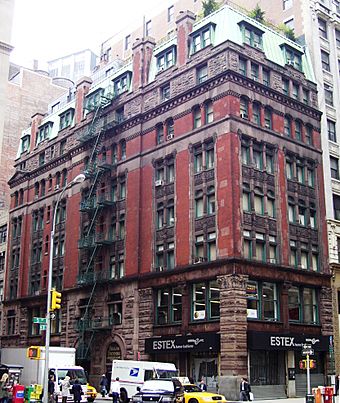 A dark red brick apartment building, with brown stone facing the lower two of its seven stories, topped by a green mansard roof with dormer windows, seen from the opposite corner of its intersection. Taller buildings are beyond it, and at street level is a storefront with "Estex" written above it.