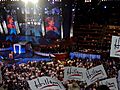 Clinton speaking on behalf of Barack Obama before a convention audience during the second night of the 2008 Democratic National Convention in Denver. Multiple audience members in the foreground wave white flags with the word "Hillary" written in marker.