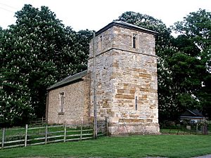 A simple stone church showing a stubby tower with pyramidal roof and lancet windows and beyond that, the nave with a single window