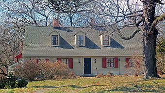 A light yellow one-and-a-half-story wooden house seen from its front, with a large bare tree obstructing part of the view at upper right. It has reddish-orange shutters on the windows and three dormer windows in its shingled roof.