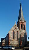 Side view of a long flint-built church with several parts, each with a different roofline.  A spire is visible behind the body of the church.  All windows are framed by brickwork.