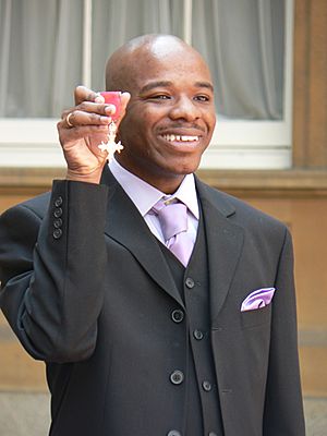 Wiltshire holding his MBE high in his right hand. He is shown from the waist up, smiling and formally dressed (black suit and waistcoat; white shirt with lilac tie, loosely tied). His head is shaved; a ring is visible on his right little finger