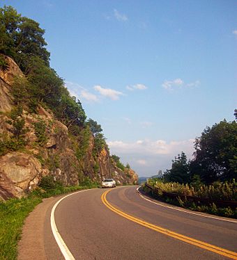 A view down a curved section of Bear Mountain Bridge Road, with a steep rock face on one side, stone wall on the other and mountains in the distance.