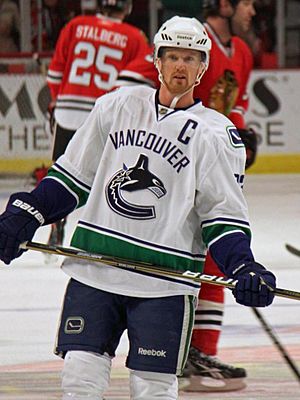 An ice hockey player wearing a white and blue jersey with a logo of a stylized orca in the shape of a "C". He stands relaxed on the ice looking forwards.